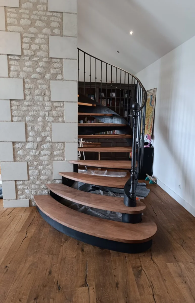 Escalier hélicoïdal aux marches de chêne et rampe en fer forgé à barreaux fins, contre un mur de pierre apparente