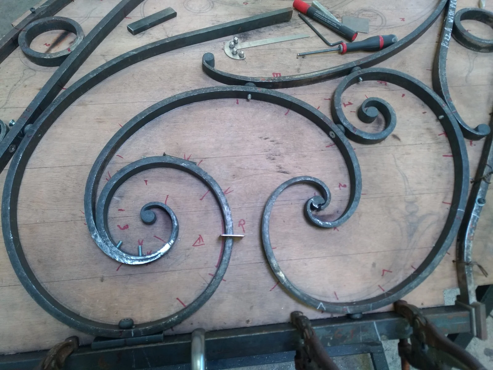Volutes de fer forgé posées sur la table de traçage, ajustées sur le dessin grandeur nature aux repères rouges